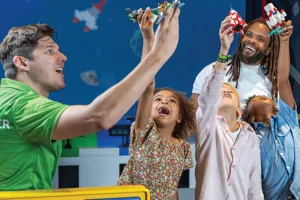 A master model builder at LEGO Discovery Center Atlanta is helping a dad and his three kids build a spaceship as part of the "Build Your Spaceship Scan and Play" attraction. The group is engaged in assembling the spaceship with colorful LEGO bricks, with the dad and kids excitedly participating in the hands-on activity. The attraction features interactive elements where participants can scan their creations for added play experiences.