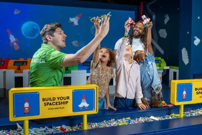 A master model builder at LEGO Discovery Center Boston is helping a dad and his three kids build a spaceship as part of the "Build Your Spaceship Scan and Play" attraction. The group is engaged in assembling the spaceship with colorful LEGO bricks, with the dad and kids excitedly participating in the hands-on activity. The attraction features interactive elements where participants can scan their creations for added play experiences.