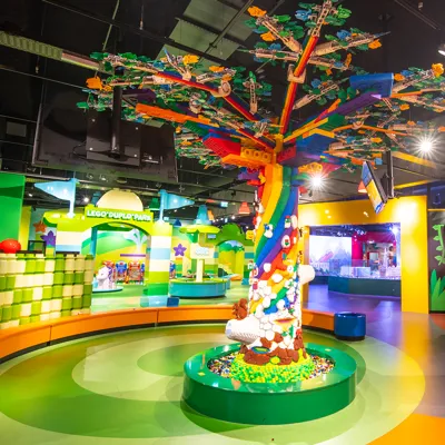 The Tree of Imagination at LEGO Discovery Center Washington DC stands tall in the center of the attraction. The large, colorful tree is made entirely of LEGO bricks, with intricate details and vibrant colors. Visitors can be seen admiring and interacting with the tree, which serves as a focal point for creative play and exploration within the center.