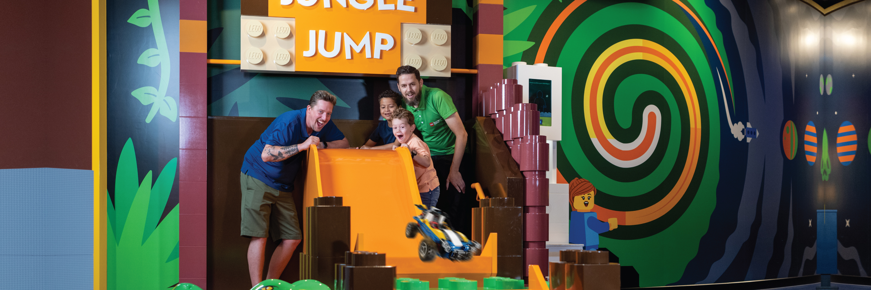 An engaging scene at the LEGO Discovery Center Washington DC shows a master model builder guiding two children as they excitedly race a colorful LEGO car down a steep ramp. The adult is kneeling beside the ramp, encouraging the kids, who are eagerly watching their creation zoom along. Surrounding them are vibrant displays of LEGO creations, highlighting the creative atmosphere of the center.